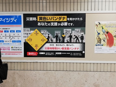 　　市内の小田急線の各駅に掲示されているポスターの写真です。大きさは　縦　約1m20�p位　横　約1m50�p位　の大きさで、そのポスターの横方向　3分の１を利用して　「災害時、黄色いバンダナ　を見かけたら　あなたの支援が必要です。」と書かれています。　左側の６分の１位のスペースにバンダナを広げた説明とその後ろに横方向　3分の1のスペースに街なかのビルなどのイラストの前に背中にバンダナをつけた人、乳母車を押す人、子供の手を引く人、犬を連れ立っている人などの形をしたイラストが書かれています。また下側3分の1の横方向のスペースを利用して　「SOS　知らせよう、　助けよう。　災害時等　障がい者支援バンダナ」と書かれ、町田市の障がい福祉課の電話番号なども書かれています。